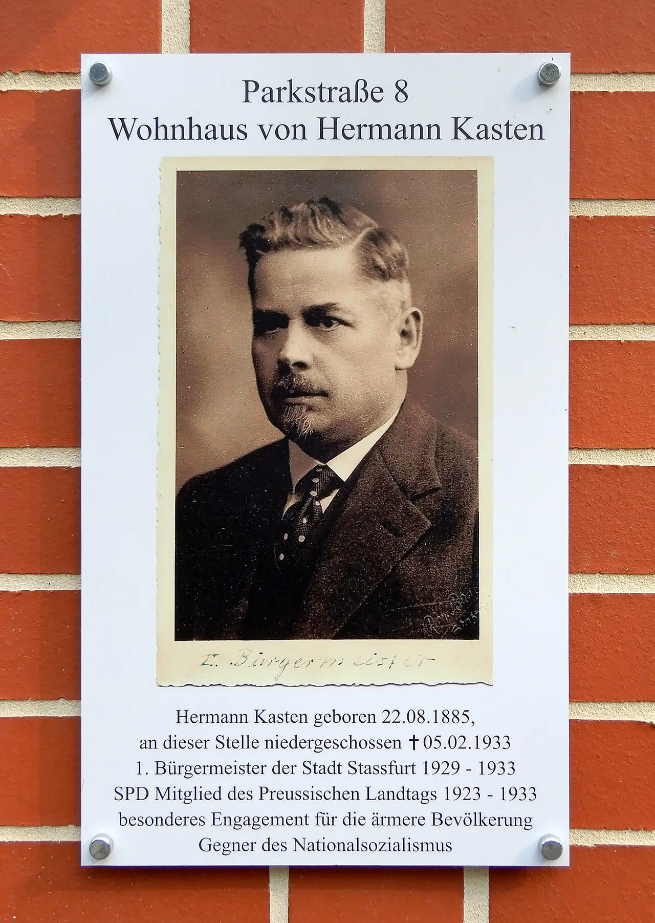 A white board on a brick wall with a sepia portrait of a moustached man in a suit and tie and a short text below