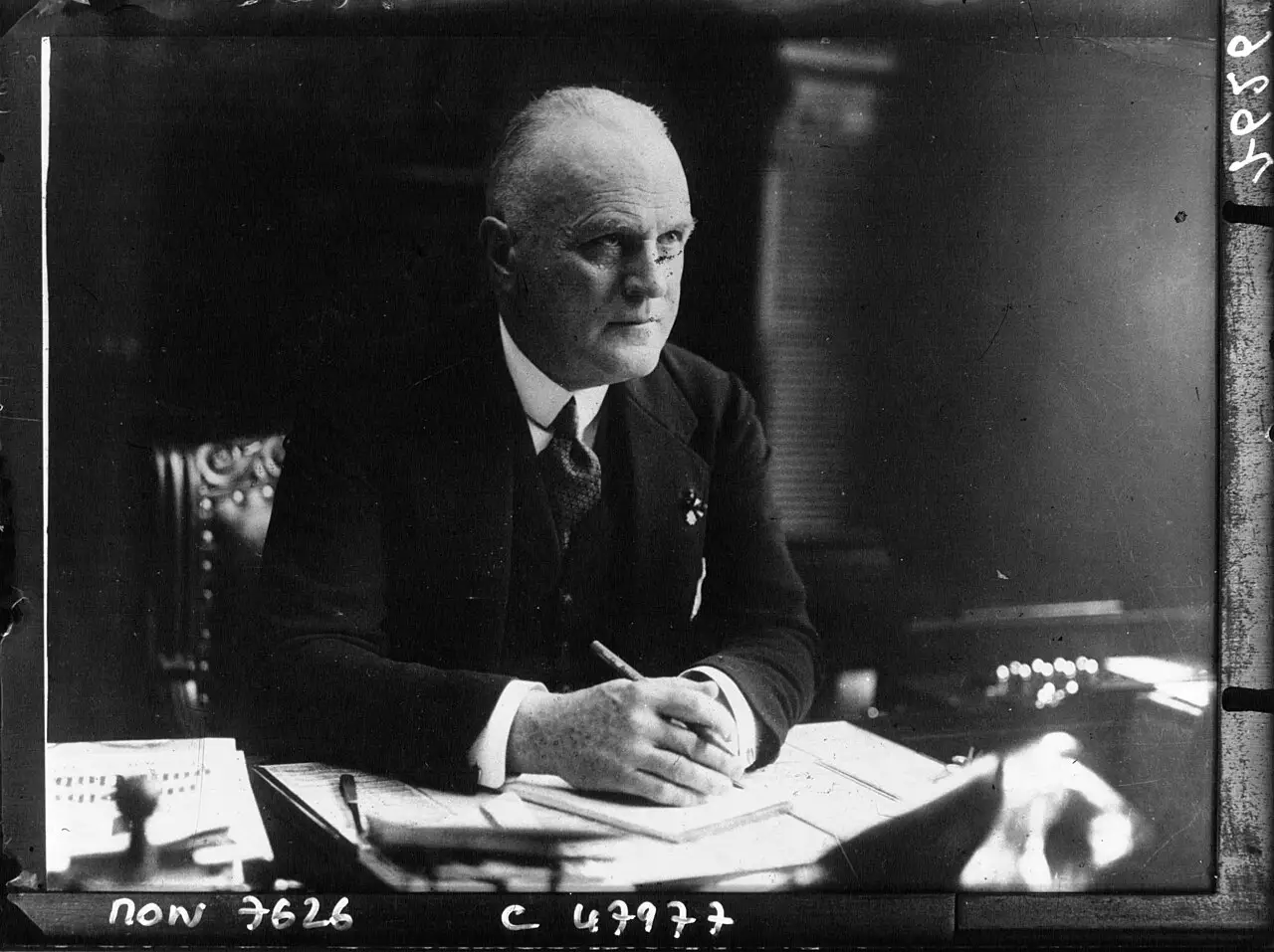 A bald, elderly man in a dark suit sits at a desk covered with papers, pen in hand, looking towards the camera.