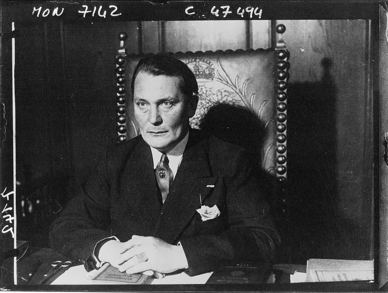 A heavy-set man in a dark suit seated at a desk in a high-backed carved chair, hands folded.