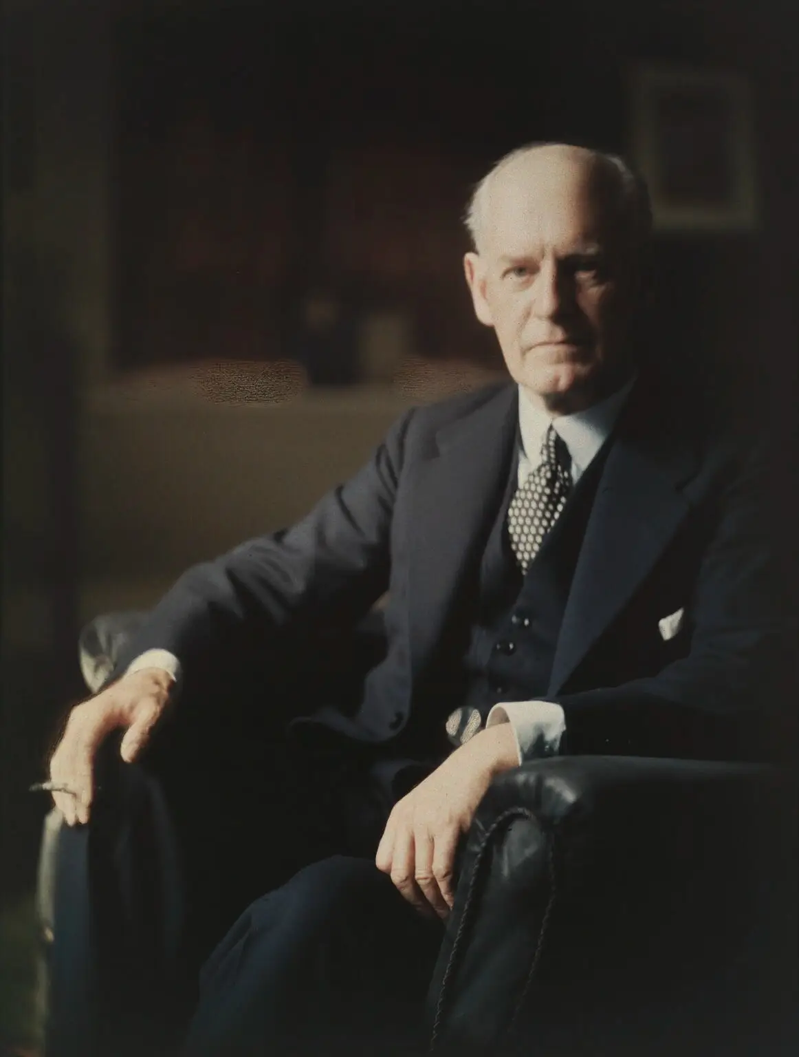 Colour portrait of a balding grey-haired man in a dark three-piece suit and spotted tie, seated in a leather armchair