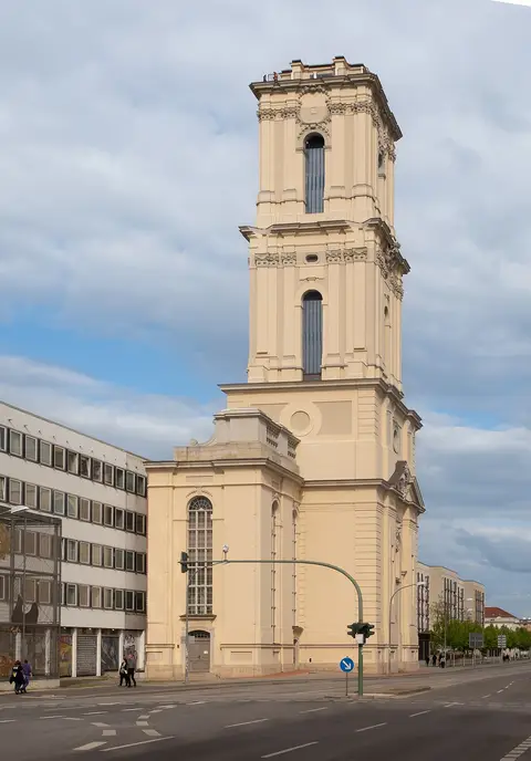 A tall pale baroque church tower with tiered upper stages against a blue sky, beside a modern building.