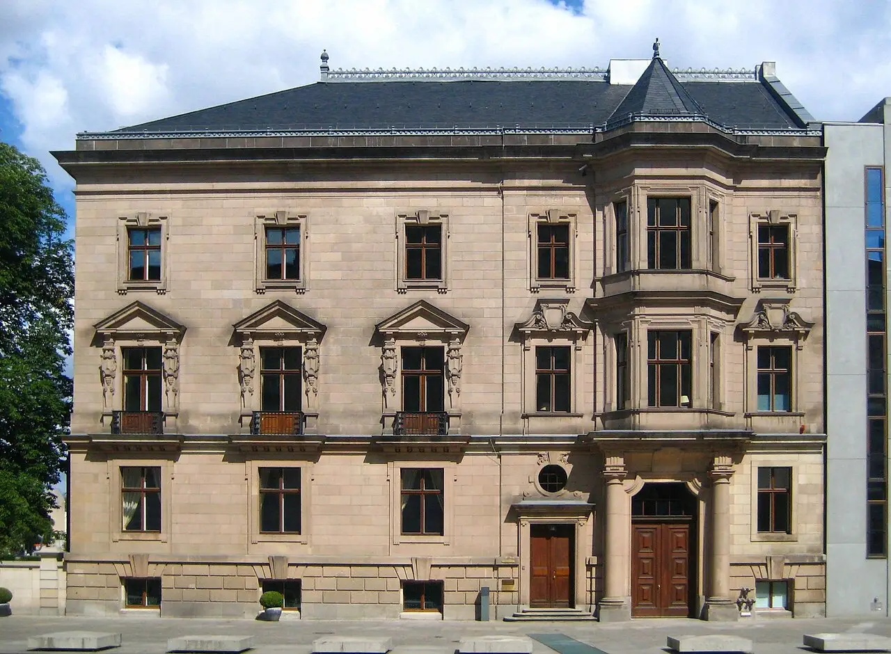 A red sandstone neo-baroque palace with a mansard roof behind railings and trees.