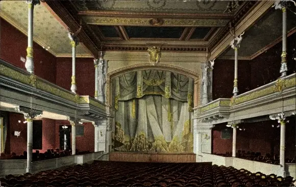 A theatre auditorium with rows of seats, two tiers of balconies on slender columns and a stage framed by a painted proscenium and curtain.