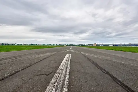 A long cracked concrete runway with a worn white centre line stretches towards a flat horizon of grass and distant buildings under a grey, cloudy sky.
