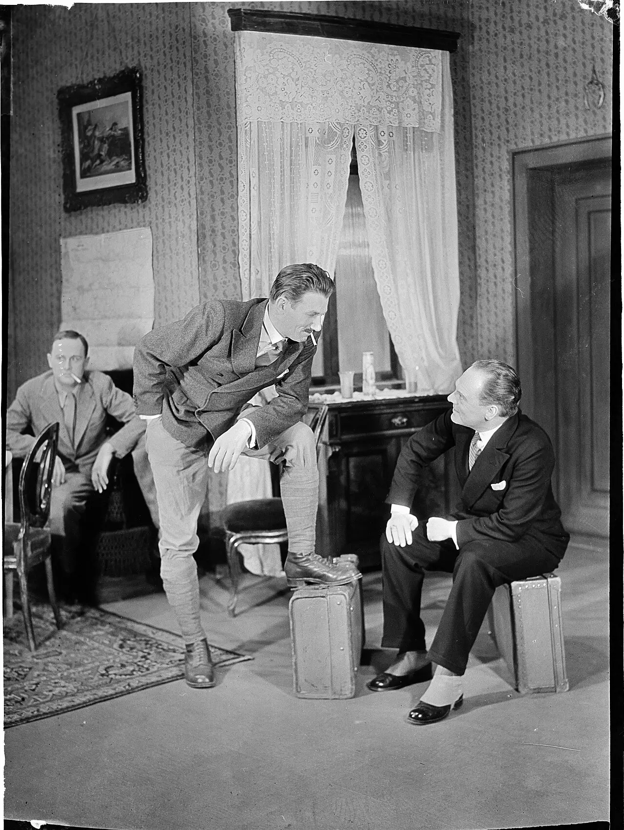 Three men on a stage set of a furnished room: one leans forward with a foot on a suitcase, another sits on a suitcase, a third watches from a chair