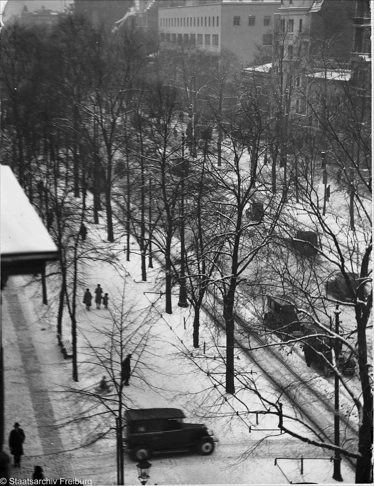 Looking down from a high window onto a snowy avenue lined with bare trees, with tram tracks, a parked car and a few people walking