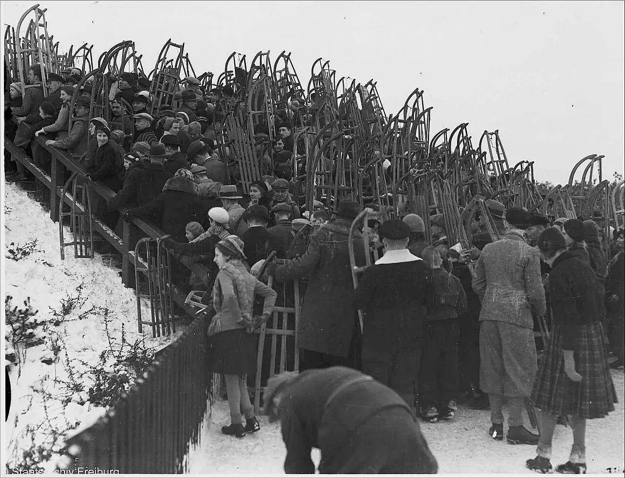 A crowd in winter coats and caps, many holding wooden sledges upright, climbs steps beside a fence on a snowy slope