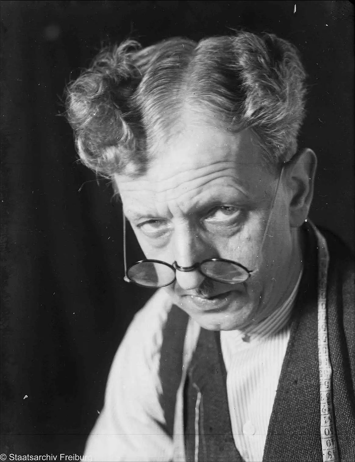 Close portrait of a grey-haired man with round spectacles low on his nose and a tape measure hanging round his neck