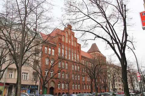 A tall red-brick school building with stepped gables and a corner tower, behind bare street trees