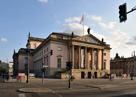 A pink-and-sandstone neoclassical opera house with a columned portico and statues on the roofline, seen across a street junction with traffic lights.