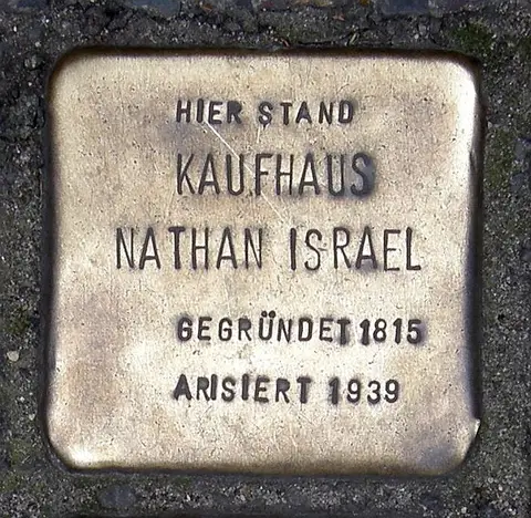 A small square brass plate set into paving stones, engraved in capitals: Hier stand Kaufhaus Nathan Israel, gegründet 1815, arisiert 1939.