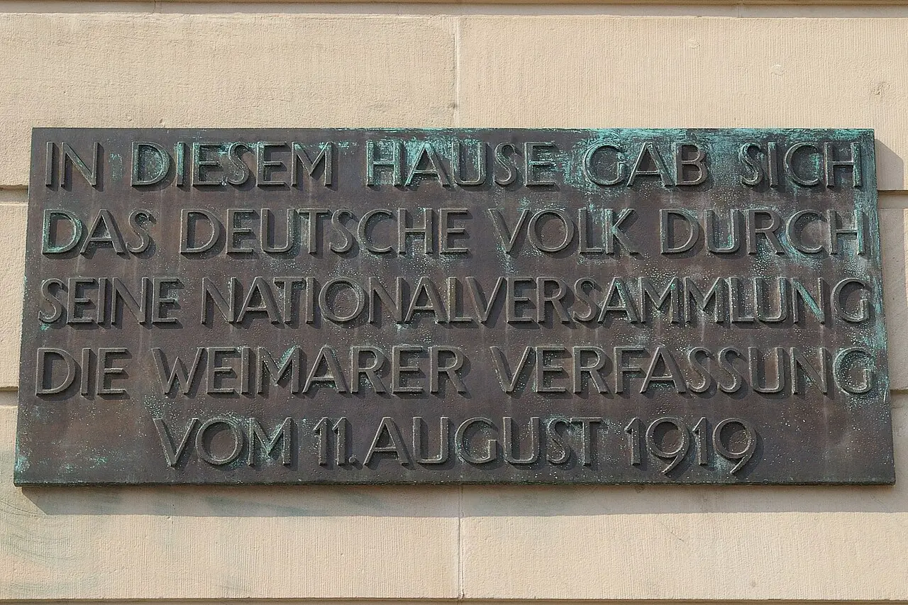 A dark bronze plaque on a pale wall with raised capital letters stating that the German people gave itself the Weimar constitution in this house.