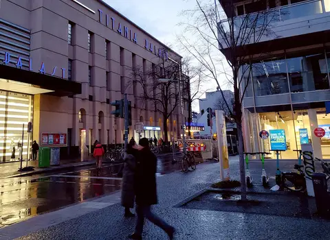 The lit lettering 'Titania Palast' on a tall rendered façade at dusk, with pedestrians, bicycles and scooters on a wet pavement.
