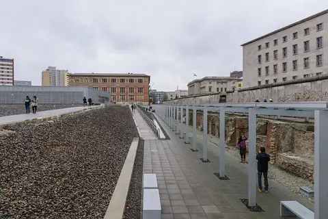 A long gravel-covered site with a walkway; on the right, old brick cellar walls under a canopy below a concrete wall, with large buildings behind