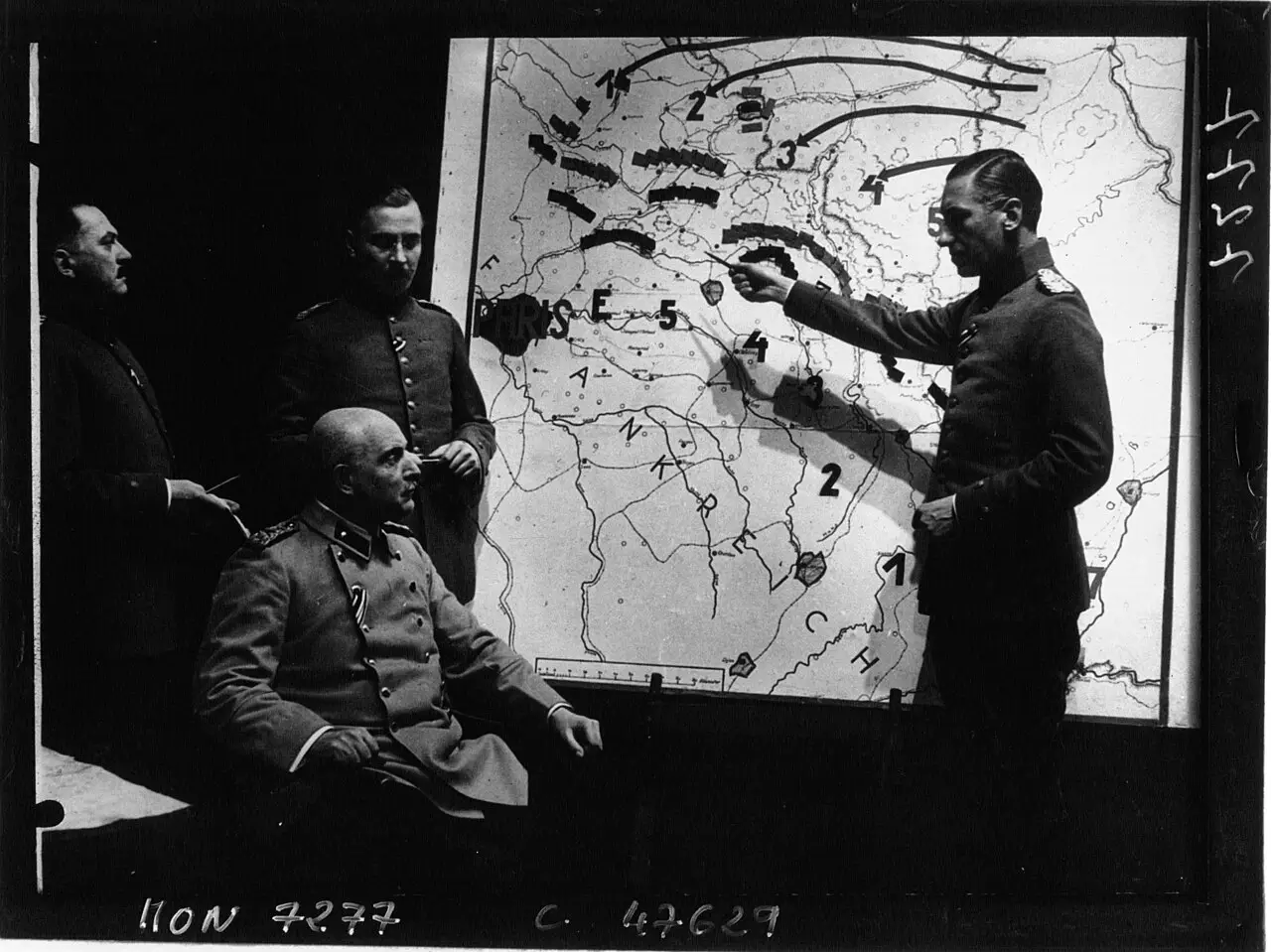Four actors in First World War German uniforms in front of a large map marked with arrows; one sits, another points at the map