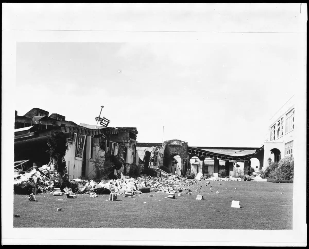 Collapsed single-storey school buildings with rubble strewn across the lawn and a damaged arcade behind.