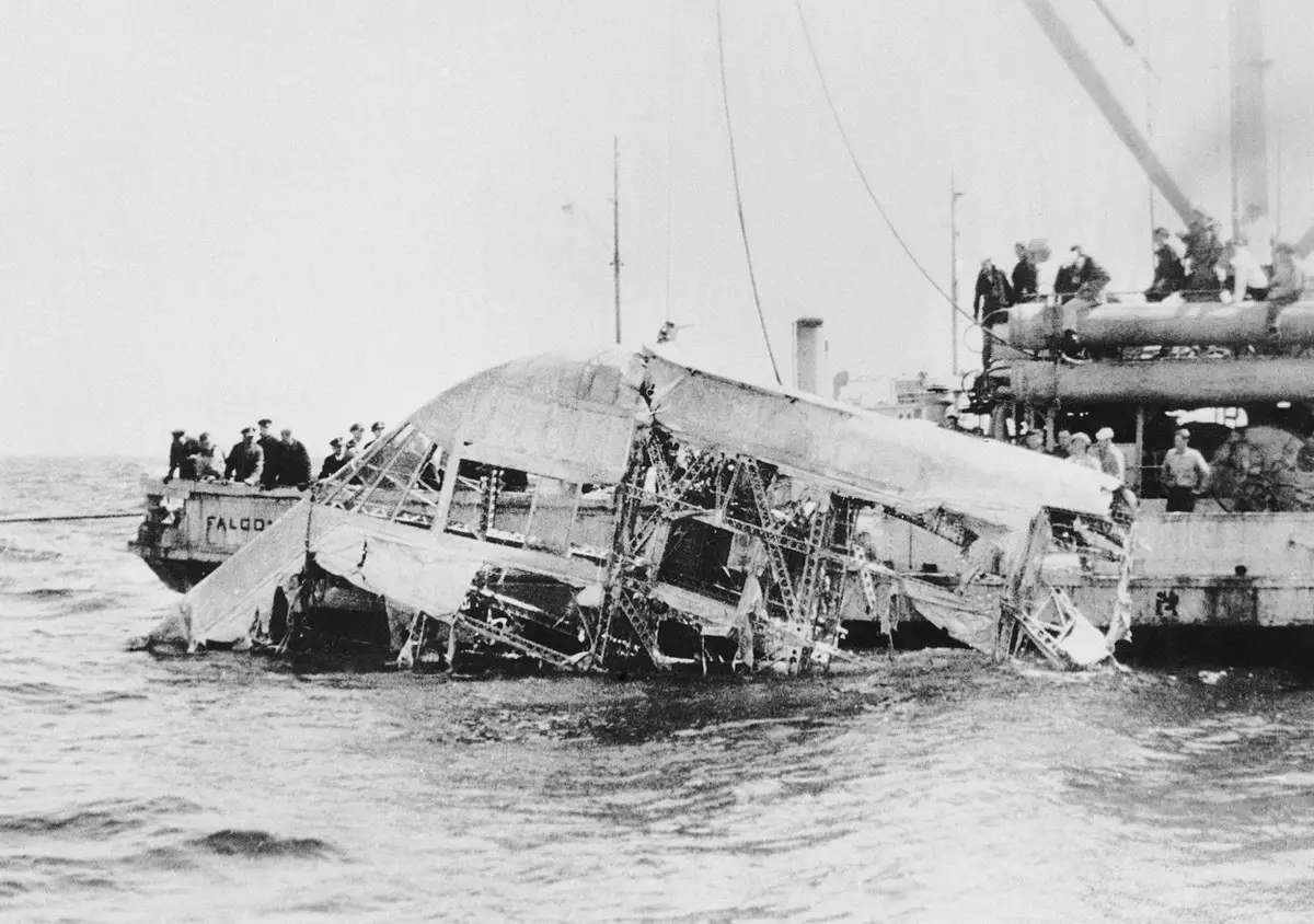 The twisted metal framework of an airship section lies half-submerged in the sea between two salvage vessels, with sailors watching from their decks.