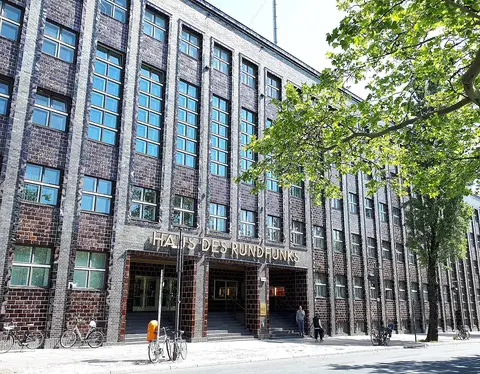 A long dark-brick building with tall vertical bands of windows and the lettering 'Haus des Rundfunks' over its entrance; bicycles stand on the pavement and a tree overhangs at right.