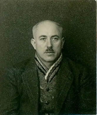 Head-and-shoulders portrait of a balding man with a moustache, wearing a jacket and a patterned scarf.