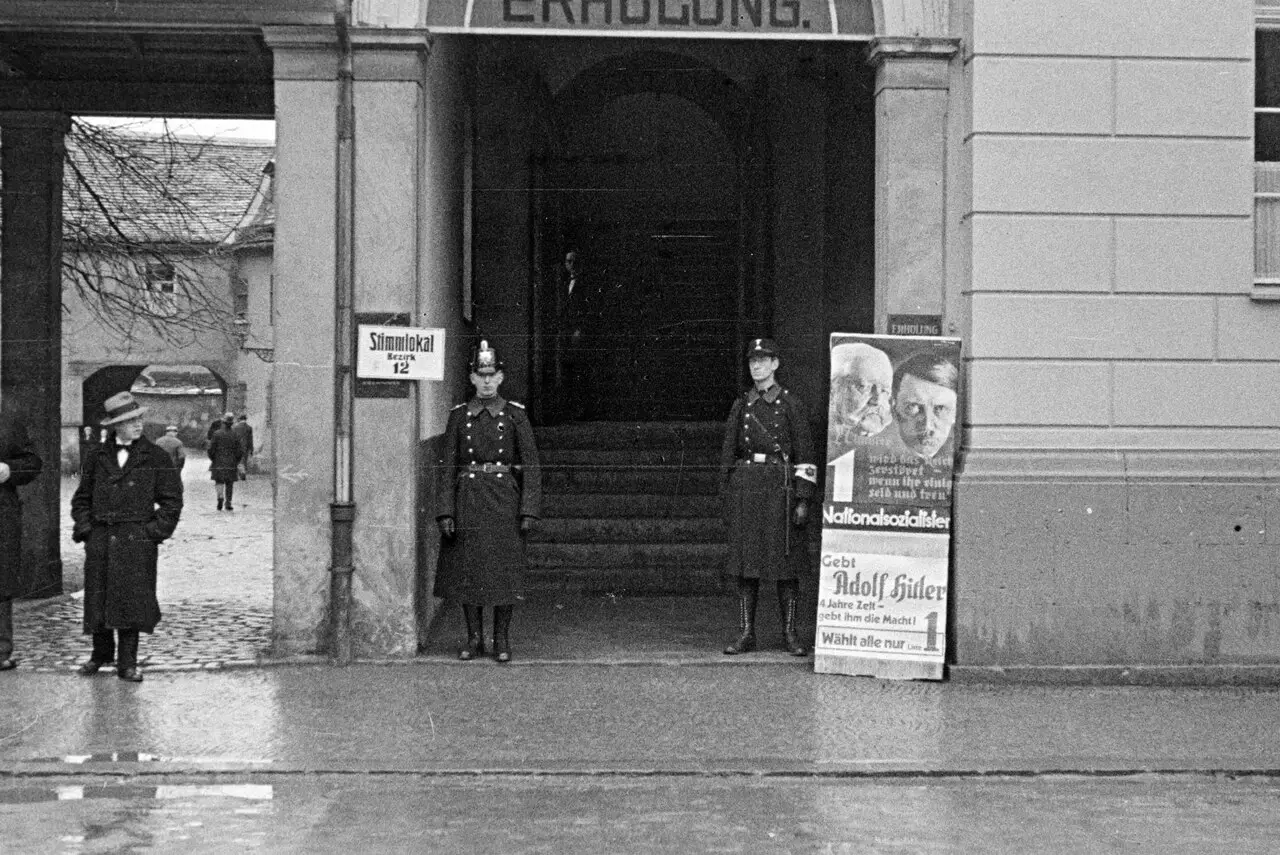 A stone gateway marked 'Stimmlokal Bezirk 12', with a police officer in a shako and a uniformed man with an armband standing either side, a poster with portraits of Hindenburg and Hitler, and a man in a bowler hat at left on a wet street.