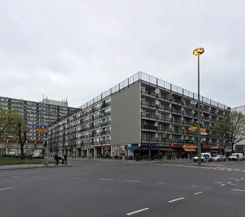 A long grey slab of flats with satellite dishes on its balconies and shops at street level, across a wide road junction